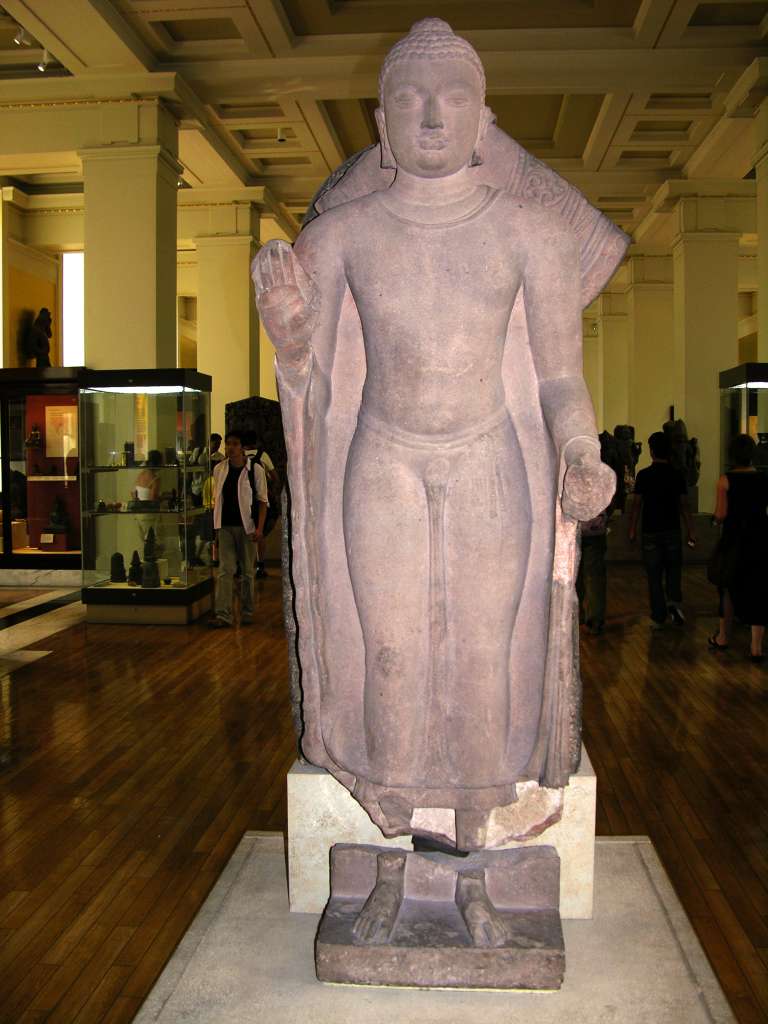 British Museum Top 20 Buddhism 06 Standing Buddha From Sarnath 6. Standing Buddha - Sarnath India, about 435AD, 1.44m high. This sandstone sculpture has heavy lidded eyes and a gentle smile marked by a pronounced lower lip, set in an oval head with heavy cheeks. This image wears a characteristically clinging garment. He holds his right hand in the fearless mudra. He has the cranial protuberance, elongated earlobes, and long arms.